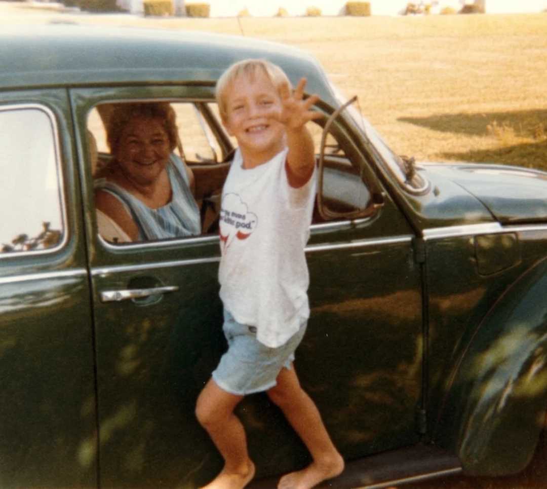 A smiling child in a white shirt and shorts stands barefoot on the door sill of an old green car, reaching toward the camera. An older woman sits inside the car, smiling at the child. It's a sunny day.
