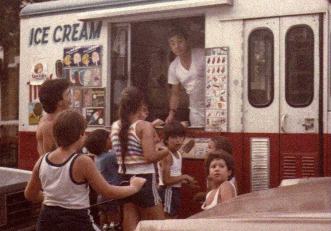 A group of children gather around an ice cream truck, looking up at the vendor inside. The truck displays colorful pictures of ice cream treats, and the scene appears to be from a past decade.