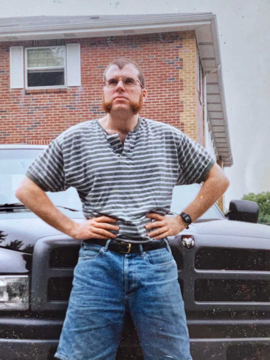 A person with unique sideburns, glasses, and a striped shirt stands confidently with hands on hips in front of a black pickup truck, with a brick house in the background.