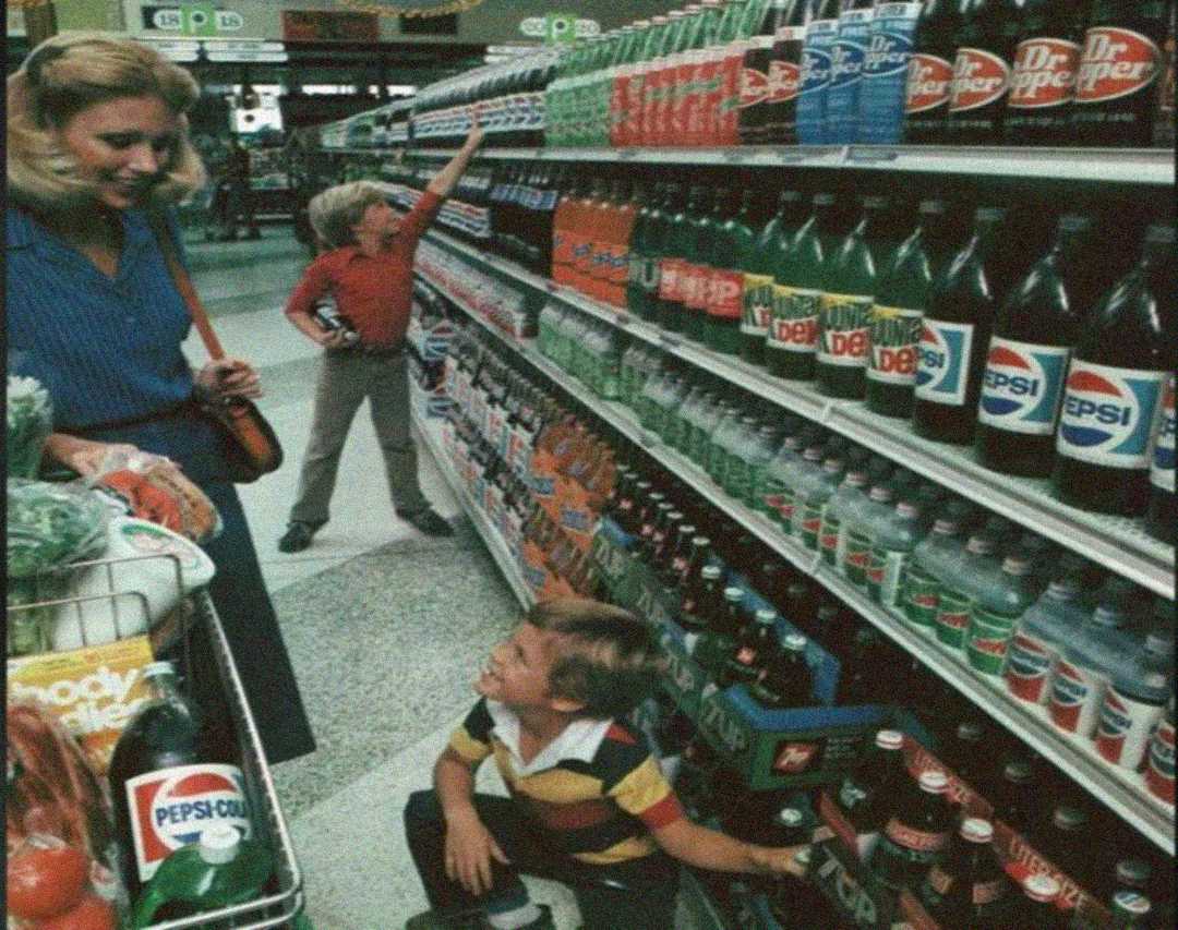 A woman with a shopping cart watches two boys in a grocery store aisle full of soda bottles; one boy is reaching up, and the other crouches while holding a bottle of Dr Pepper.