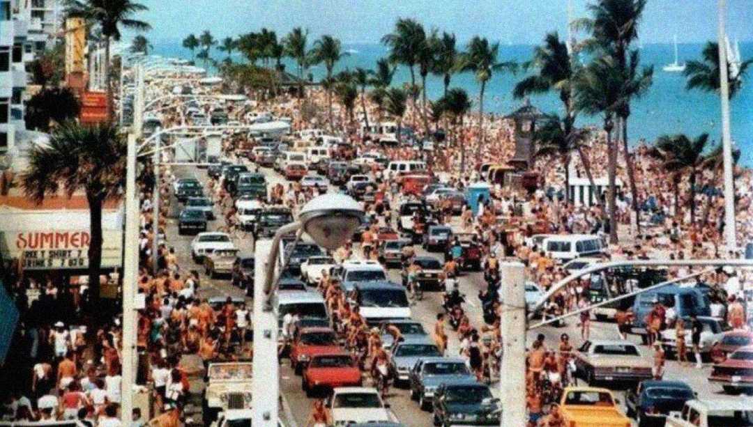 A crowded beachside street packed with cars and people; palm trees line the road and the sandy beach is filled with sunbathers and swimmers under a sunny sky.