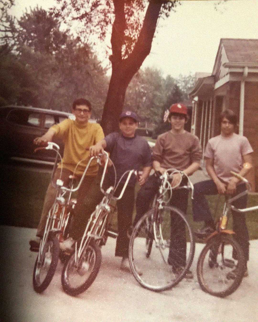 Four boys pose with their bikes on a driveway in front of a house. Trees and a parked car are visible in the background. The photo has a vintage, nostalgic feel.