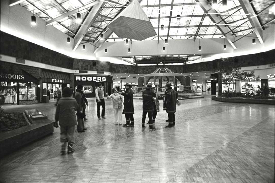 A group of people stands in a spacious shopping mall atrium with high ceilings, skylights, and stores like Rogers and Stride Rite visible. The floor is tiled, and indoor planters and benches line the open space.