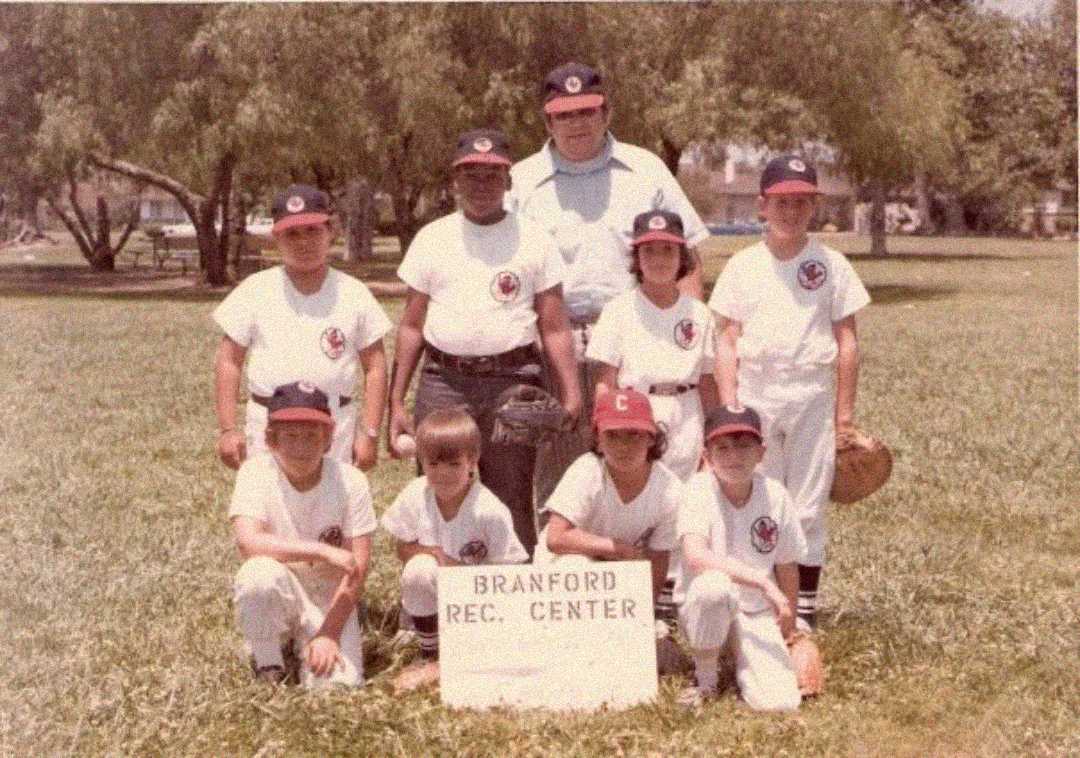 A vintage photo of eight boys in baseball uniforms and two adult coaches posing outdoors on grass with trees behind them. A sign in front reads "Branford Rec. Center.