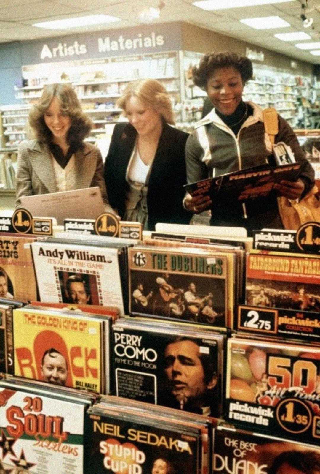 Three women smile while browsing vinyl records in a store. The shelves in front display album covers, including artists like Andy Williams, Perry Como, and Neil Sedaka. A sign above reads "Artists Materials.