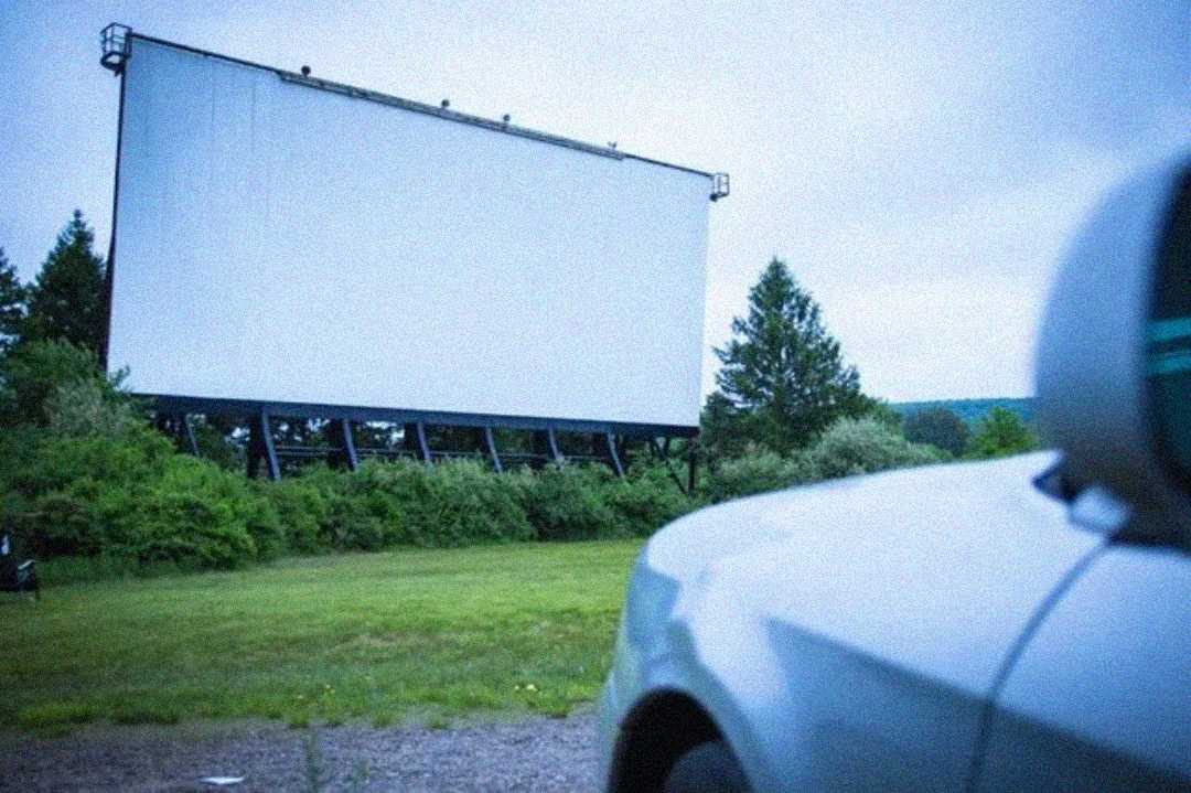 A large, empty drive-in movie screen stands outdoors, surrounded by trees and greenery, with the front of a white car visible in the foreground.