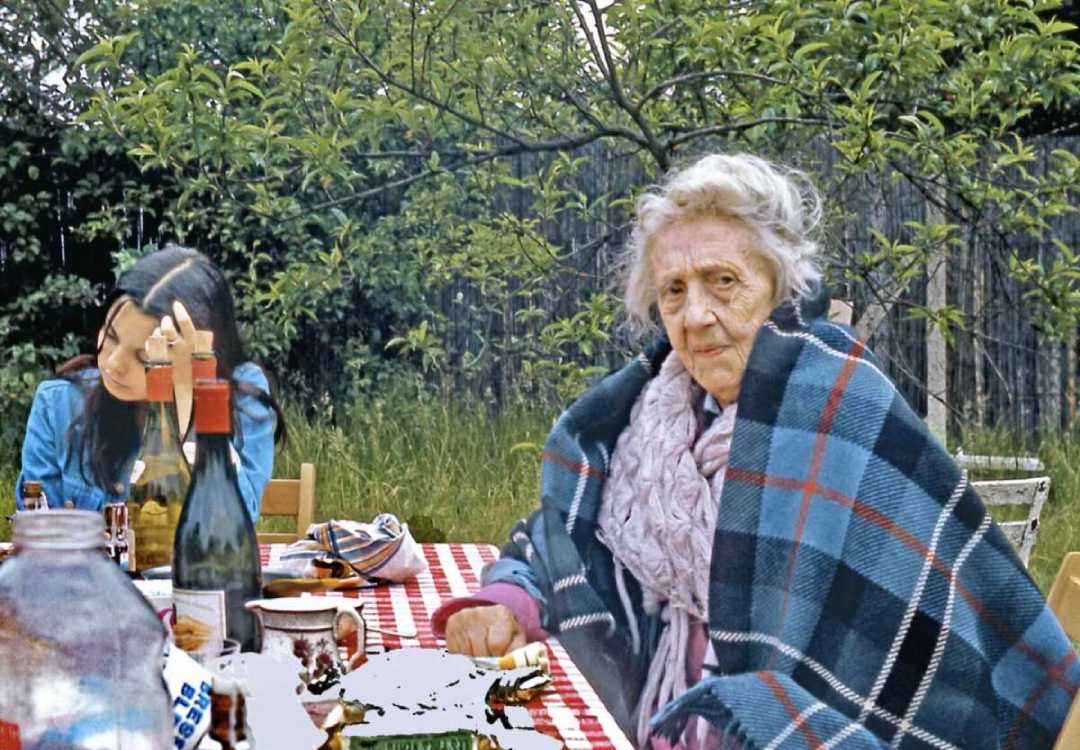An elderly woman wrapped in a plaid blanket sits at an outdoor table with a younger woman beside her. The table is covered with a red checkered cloth, dishes, and bottles. Lush green trees and grass are in the background.