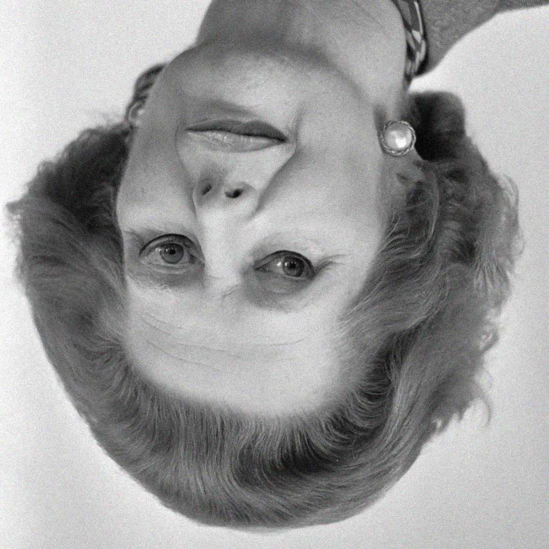 Black and white photo of an older woman with styled hair, pearl earrings, and a necklace, shown upside down.