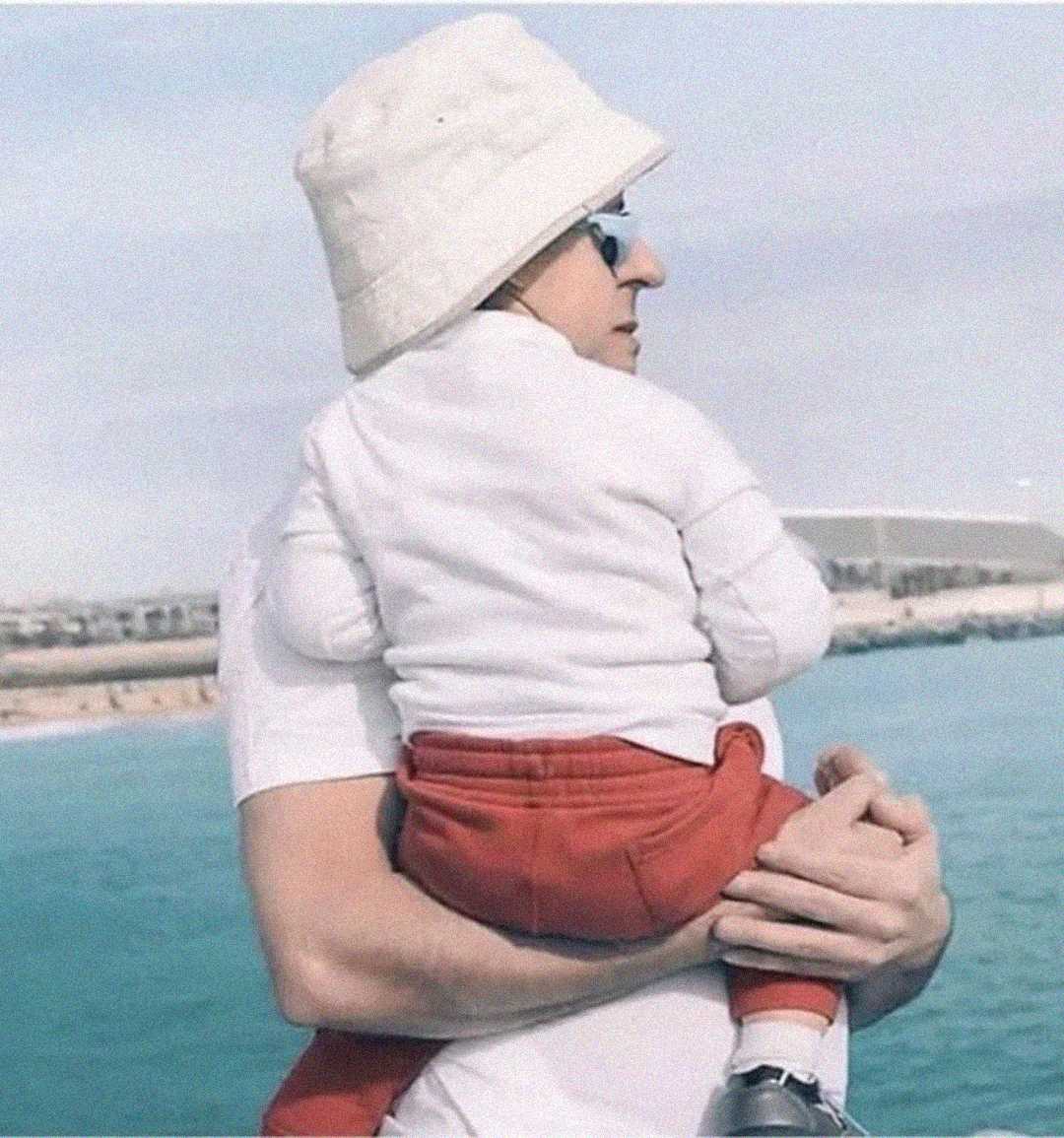 An adult wearing sunglasses and a white bucket hat holds a young child dressed in a white top and red pants by the seaside, with blue water and a distant shoreline in the background.