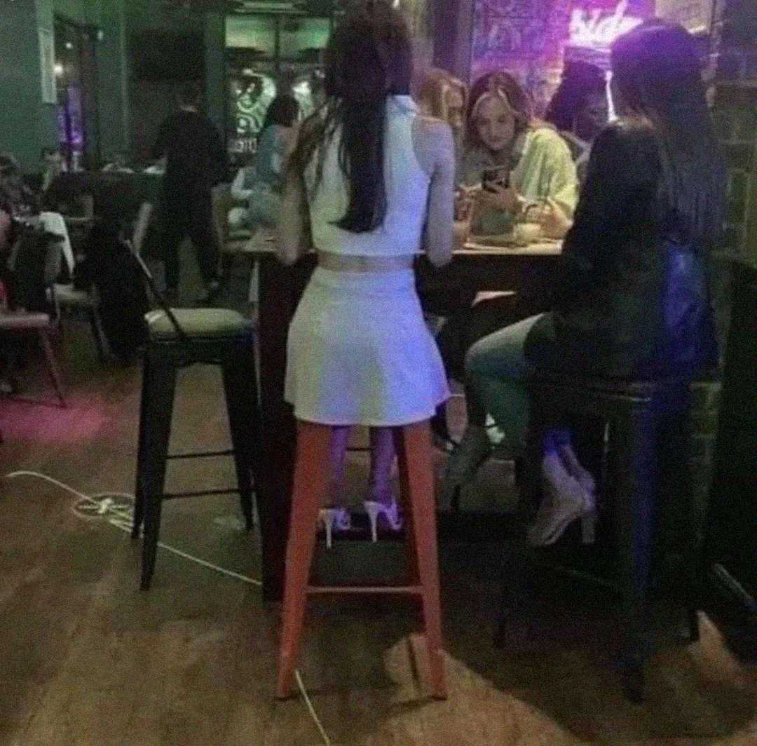 A woman in a white outfit sits on a tall barstool at a high table with three other people in a dimly lit bar or restaurant, with drinks and phones visible on the table.