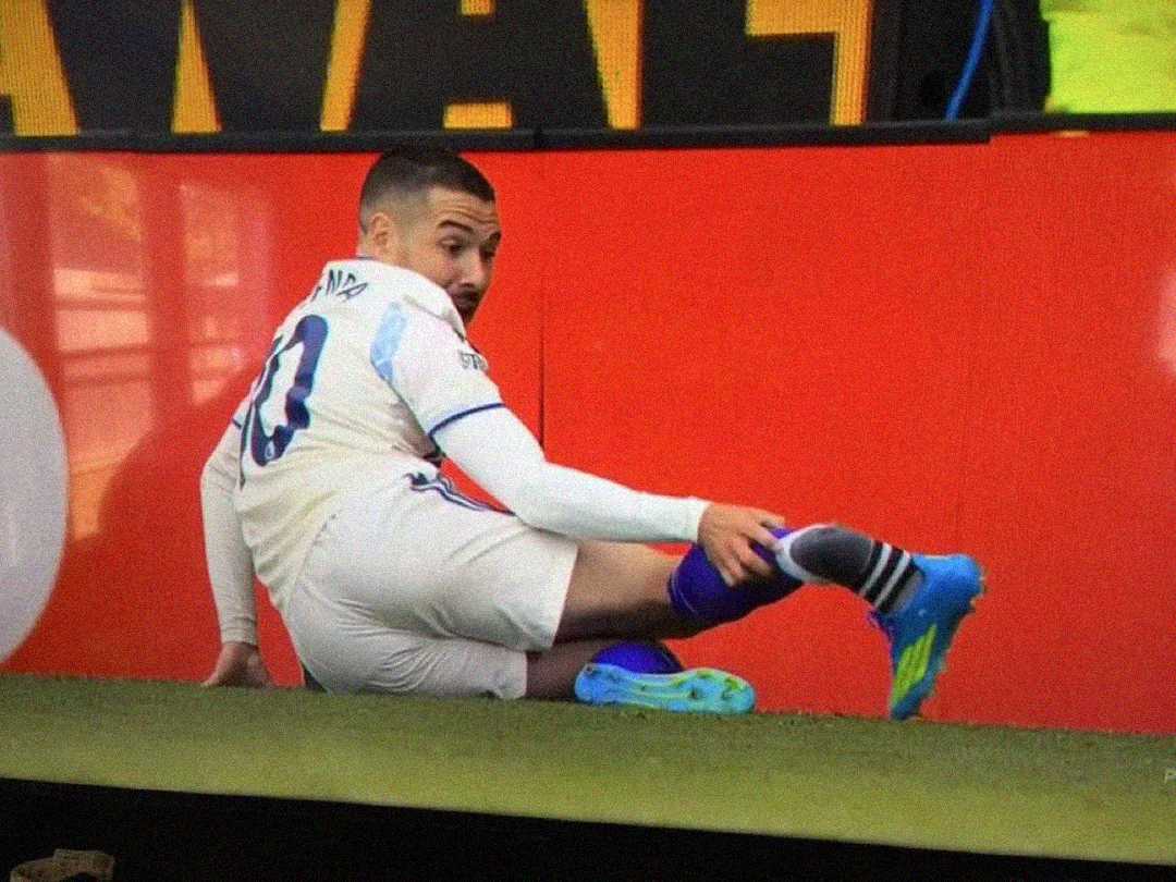 A soccer player in a white uniform sits on the field near the sideline, glancing back over his shoulder, with one leg extended and his hand touching his shin.