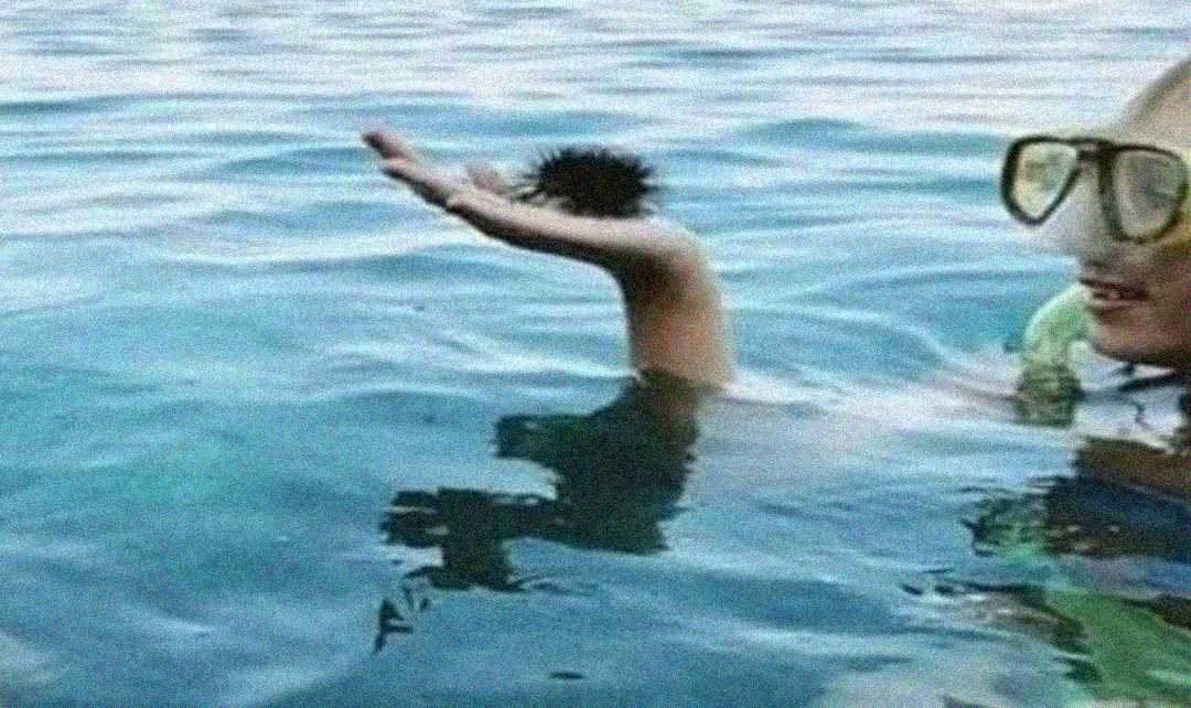 A person wearing goggles holds a black sea urchin above the surface of clear blue water while swimming.