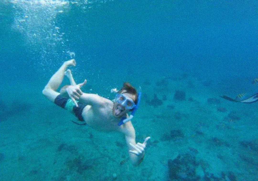 A person snorkeling underwater gives a thumbs-up and sticks out their tongue, wearing swim goggles and a snorkel. The water is clear, revealing a blue sea with rocks and some fish below.