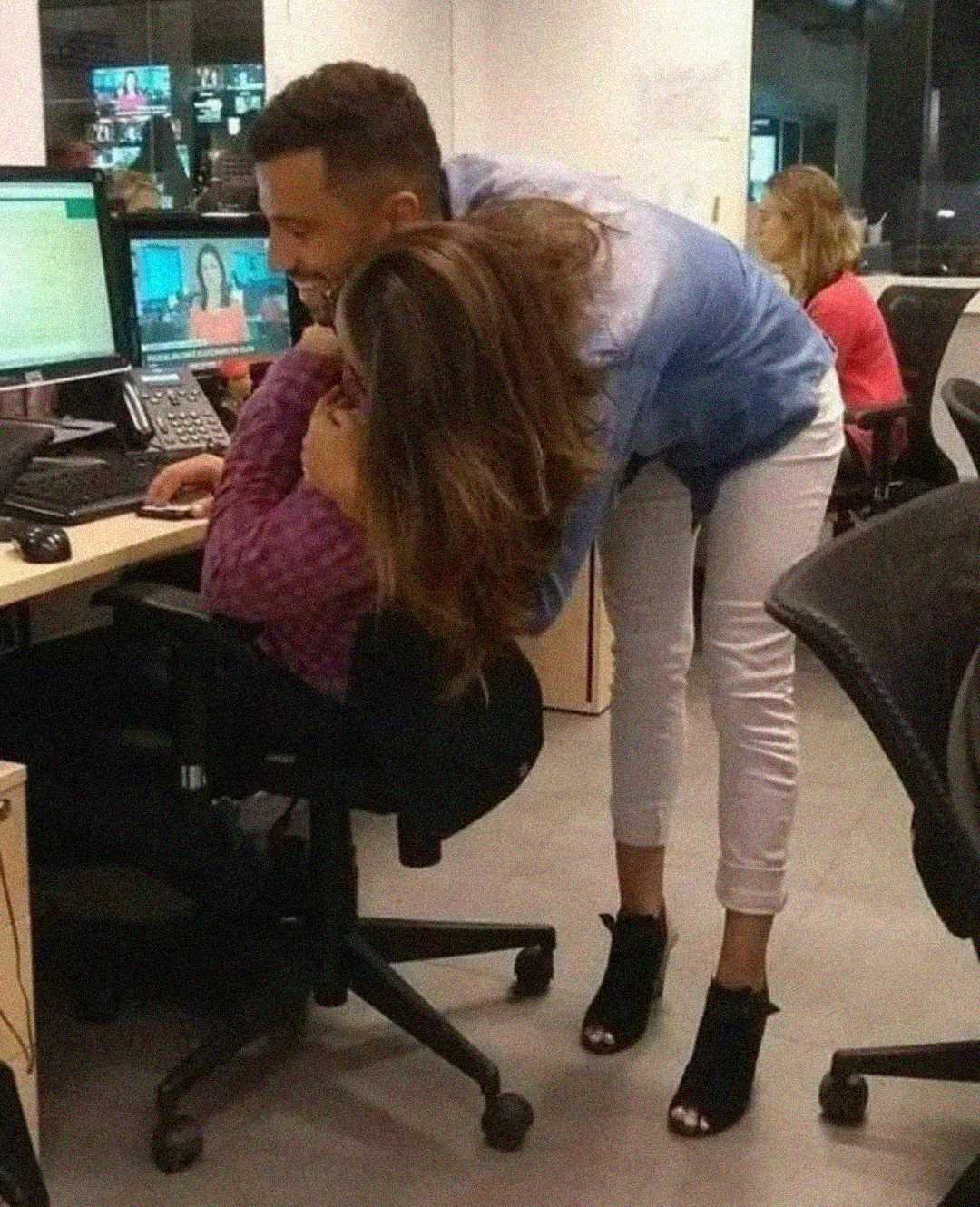 A man leans over a woman seated at an office desk, embracing her from behind as she laughs and covers her face. Another woman works at a computer in the background. The office has a casual, friendly atmosphere.
