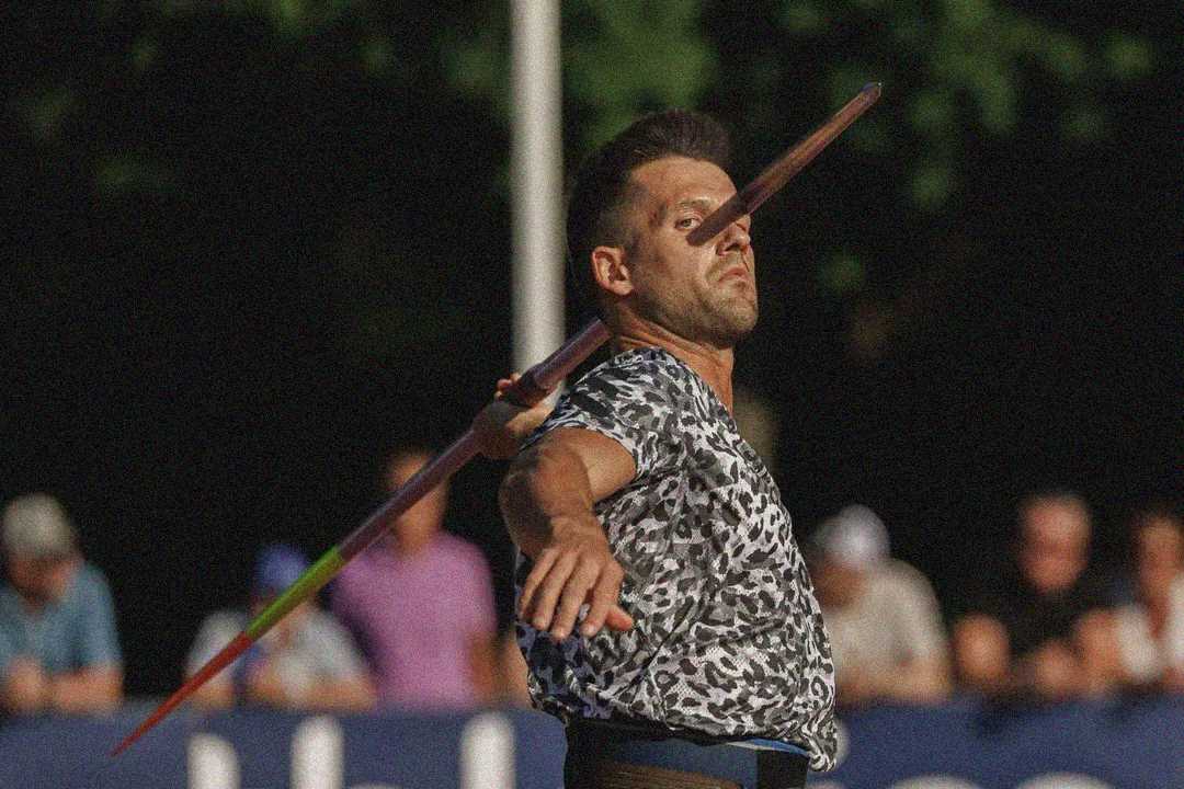 A male athlete in a patterned shirt throws a javelin, his arm extended and javelin released mid-air, with spectators blurred in the background.
