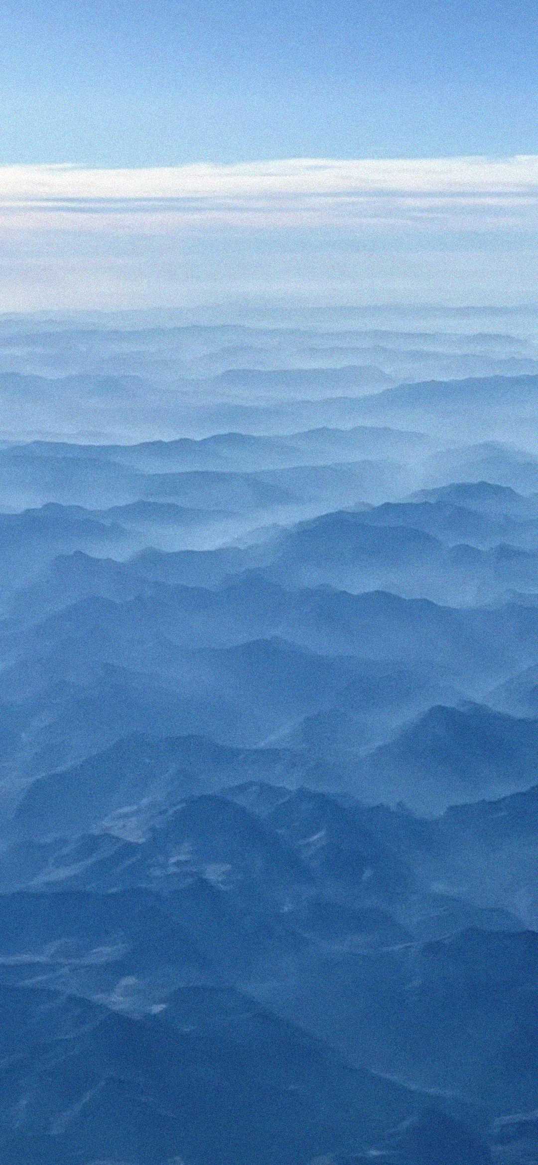 Aerial view of blue mountain ridges layered with mist, stretching into the distance under a pale sky with thin, horizontal clouds.