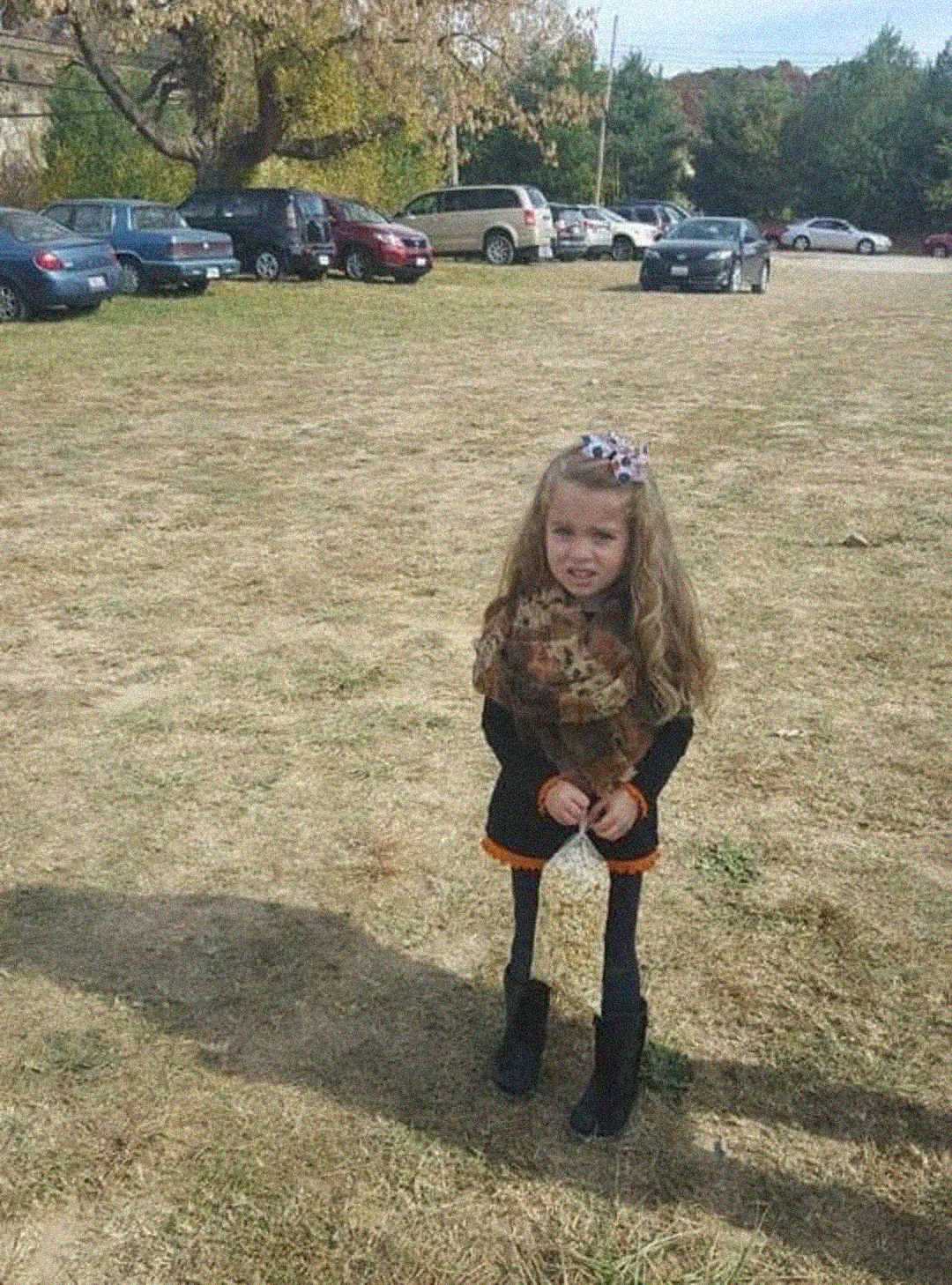 A young girl with long hair, wearing a bow, scarf, dress, and boots, stands on a grassy field with parked cars in the background on a sunny day.