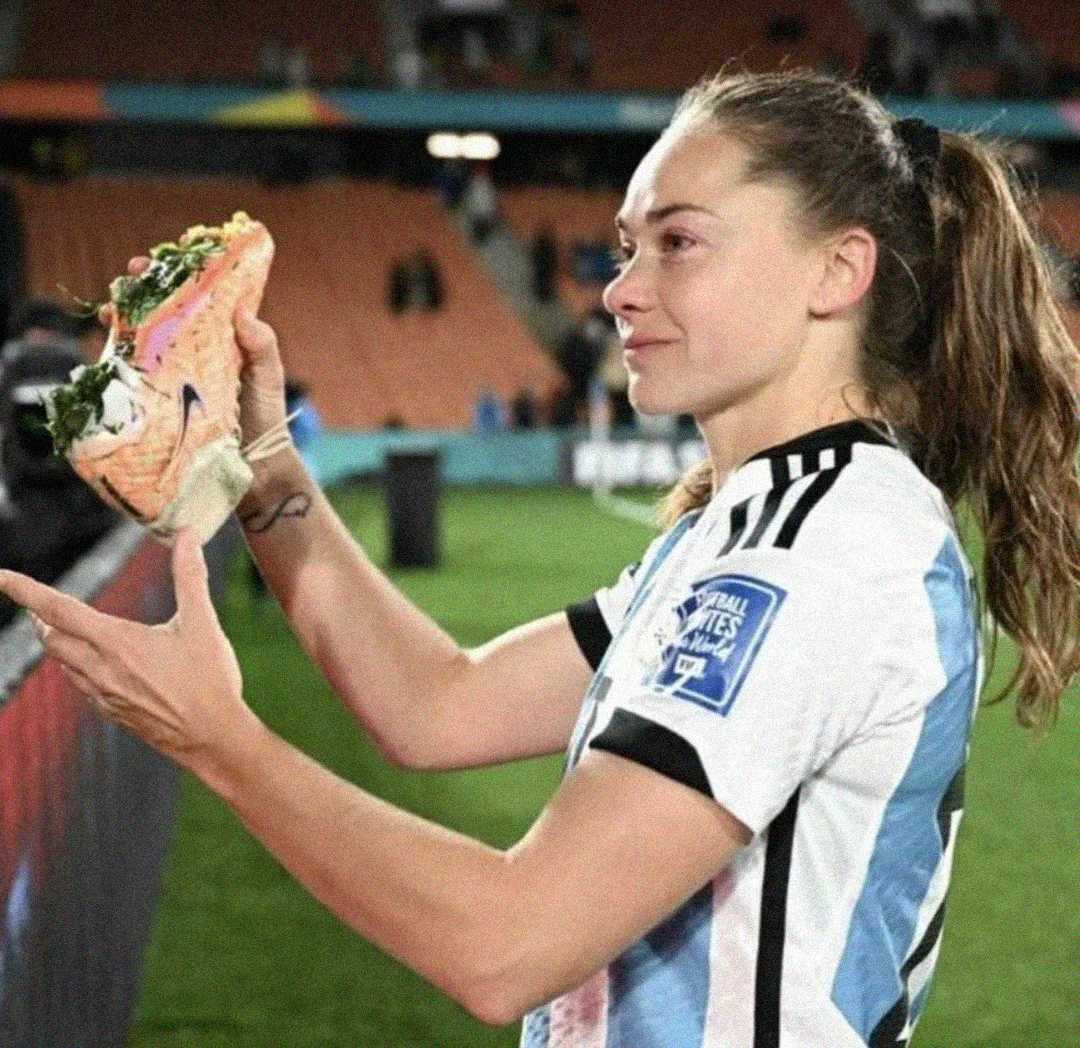 A female soccer player in an Argentina jersey holds up a large sandwich on the field, smiling, with empty stadium seats in the background.