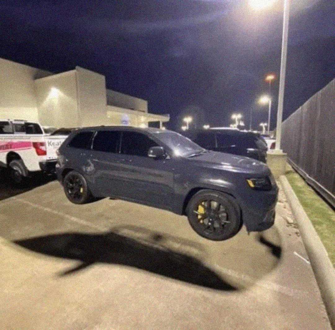 A dark SUV appears to be floating above the ground in a well-lit parking lot at night, with other vehicles and a building in the background.
