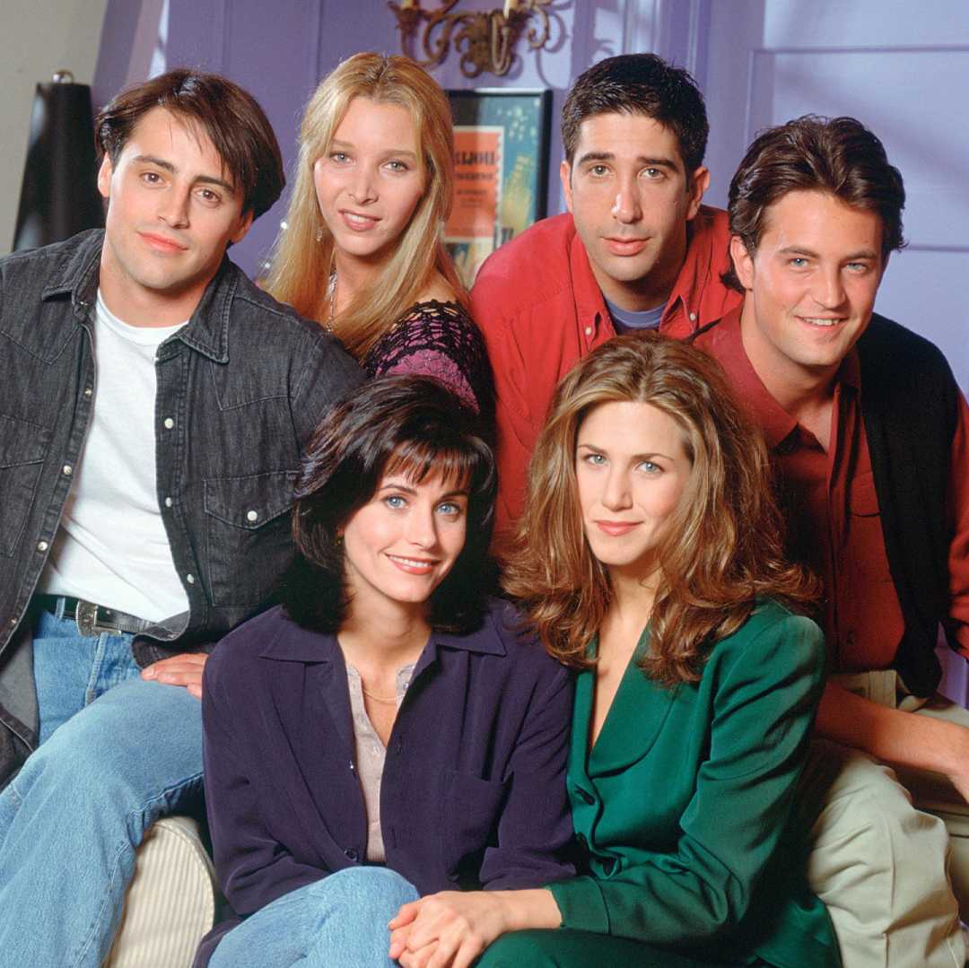 Six young adults pose closely together indoors, all smiling. They wear casual 1990s-style clothing, with a purple wall and a framed poster in the background.