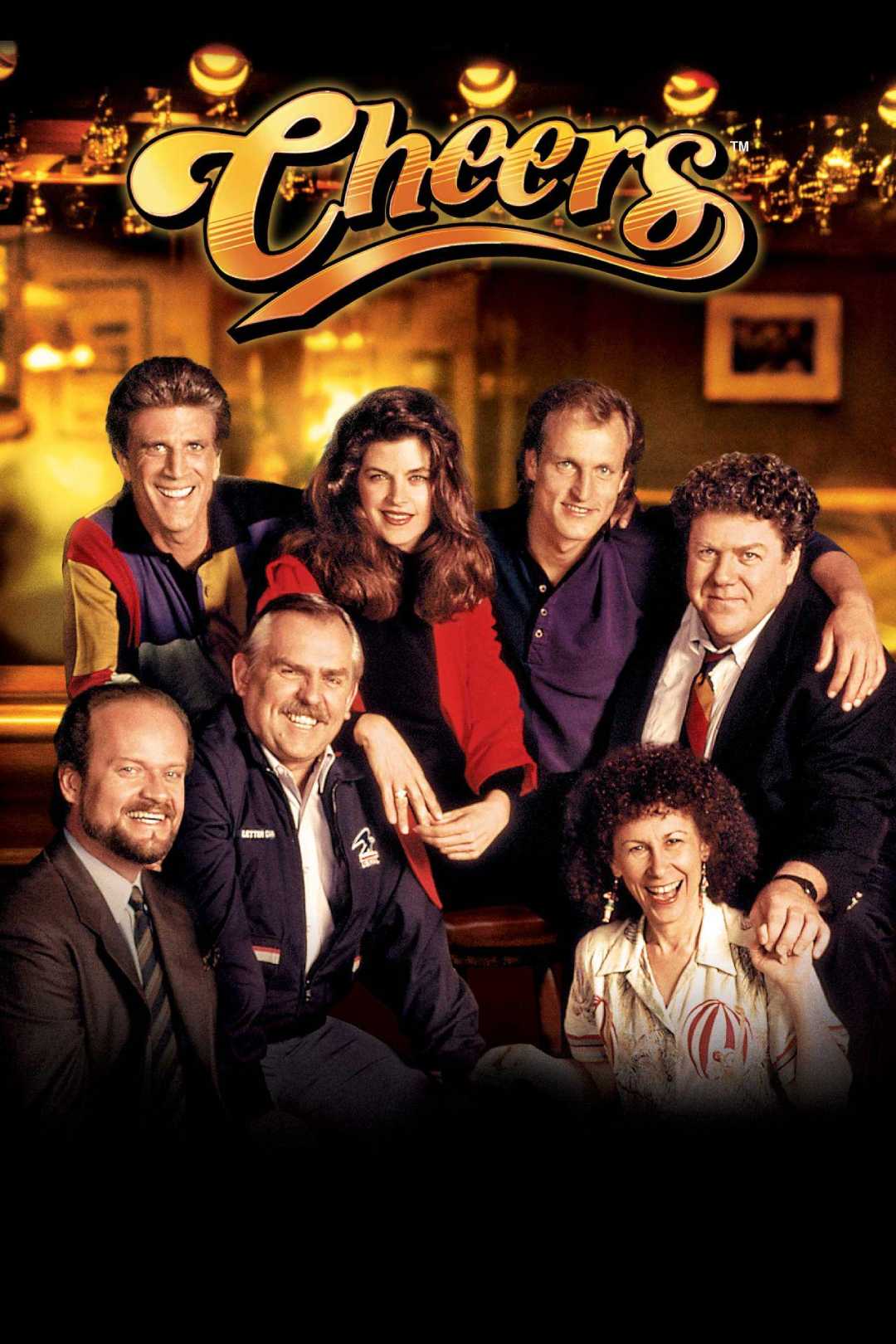 A group of seven people, smiling and sitting closely together in a cozy bar setting under the illuminated "Cheers" sign, representing the main cast of the TV show "Cheers.