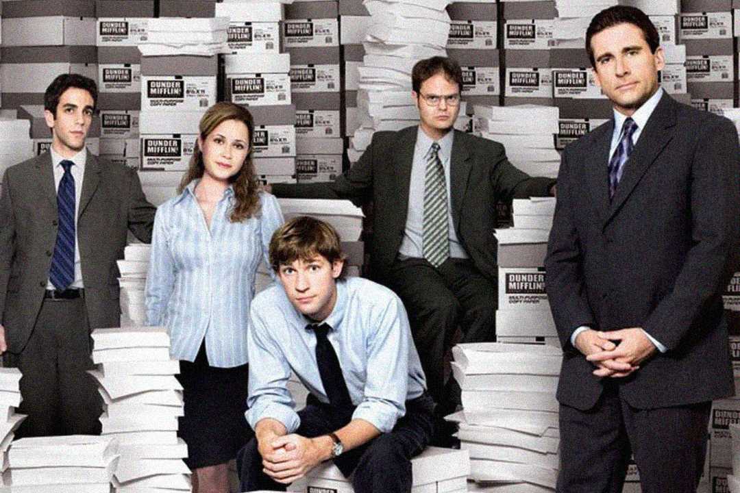 Five people in office attire pose among tall stacks of paper in an office supply warehouse setting. Boxes labeled "Dunder Mifflin" are stacked behind them, suggesting a work environment related to paper products.