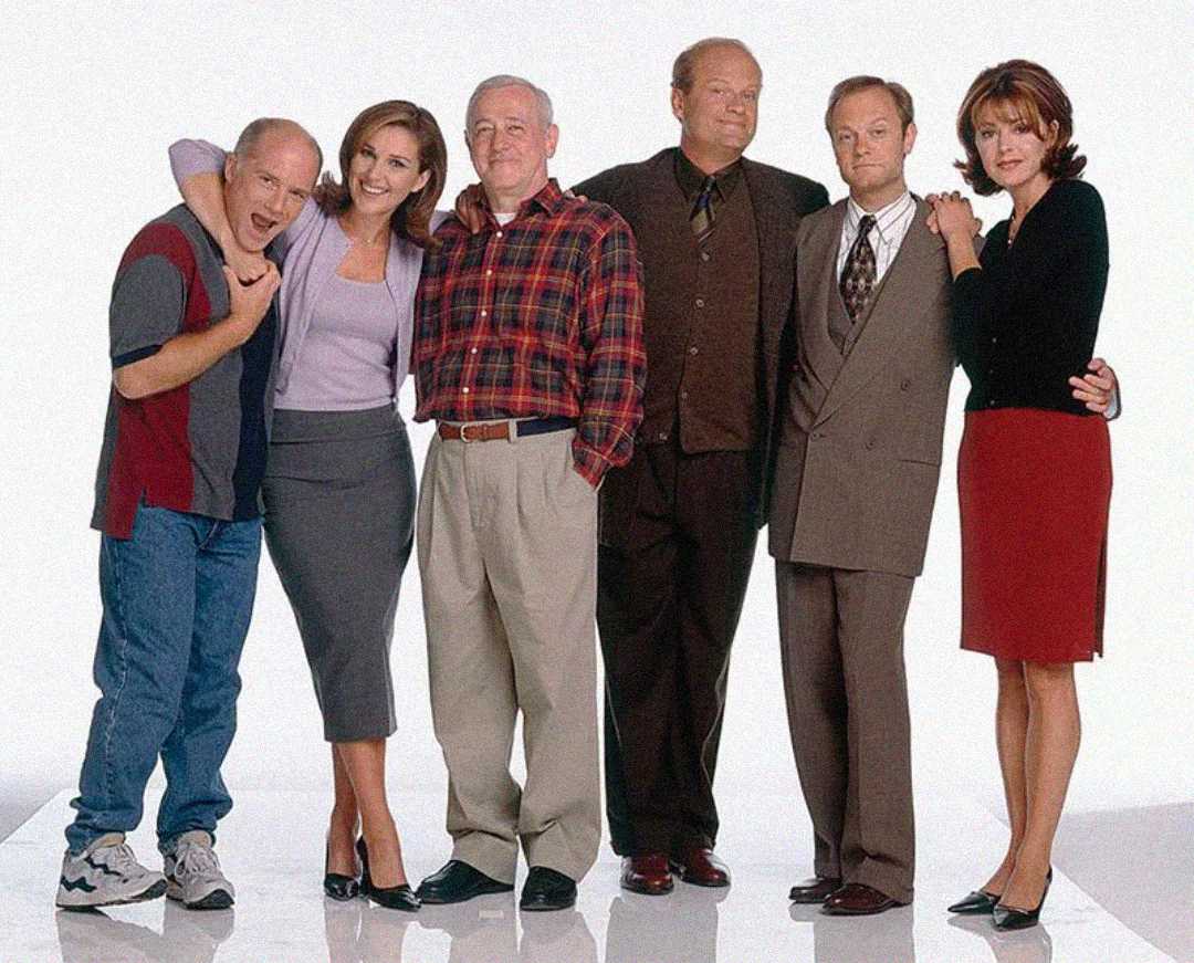 Six people stand closely together, smiling for a group photo against a white background. Their clothing is business casual, and their poses suggest friendship and camaraderie.