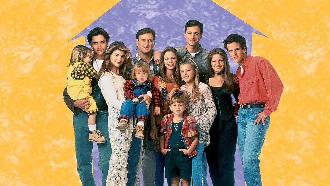 A group of adults and children stand together, smiling, in front of a yellow background with a blue house shape. They are dressed in casual 90s clothing and pose closely as if for a family portrait.