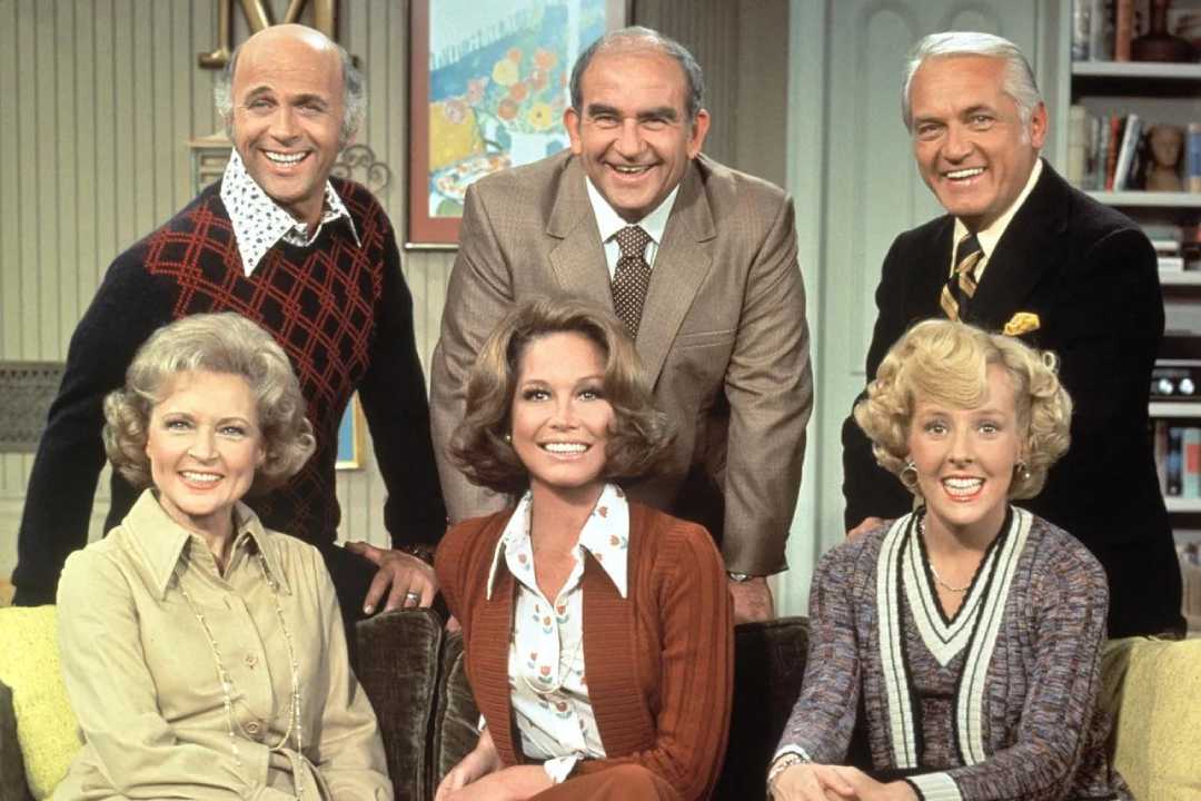 Six adults, three men standing behind three women sitting on a couch, all smiling in a warmly lit living room with framed pictures and bookshelves in the background.