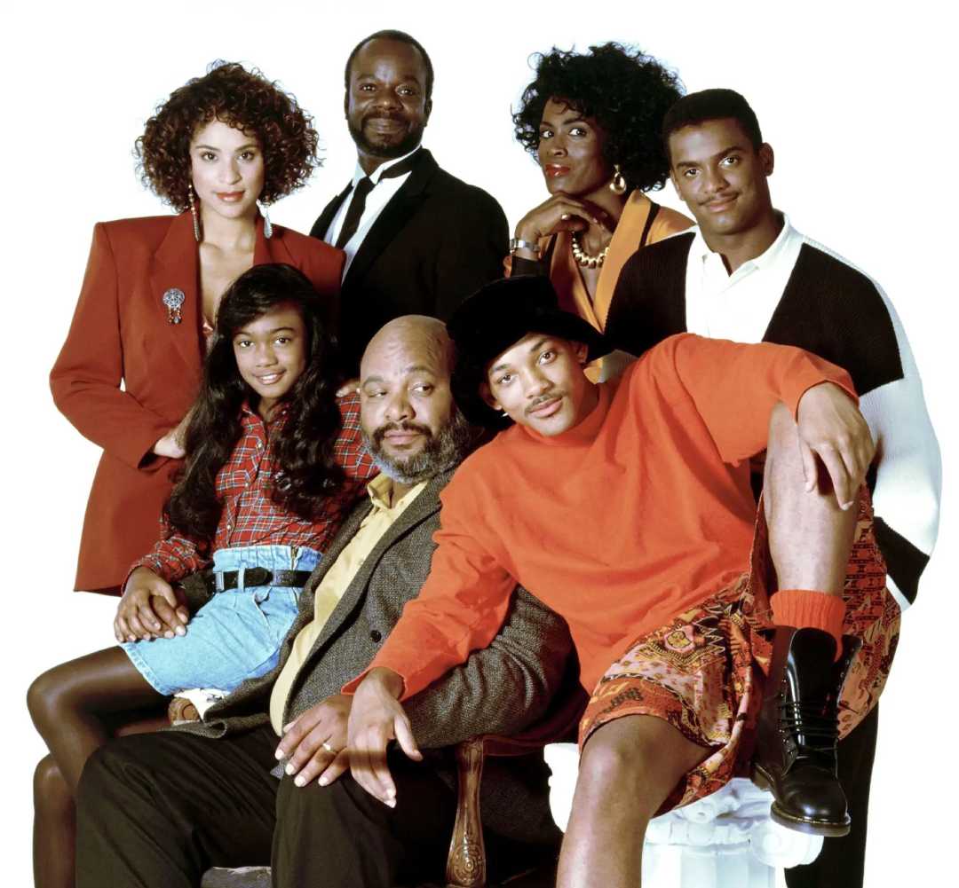 Seven people pose together against a white background, dressed in 1990s-style clothing, with some standing and others sitting, suggesting a family or close-knit group.