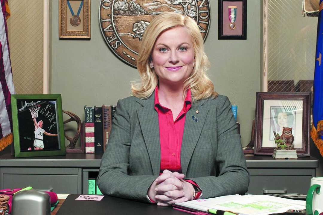 A woman with blonde hair in a gray blazer and pink shirt sits at a desk, smiling. Behind her are books, framed photos, an owl figure, medals, and the seal of the City of Pawnee on the wall.