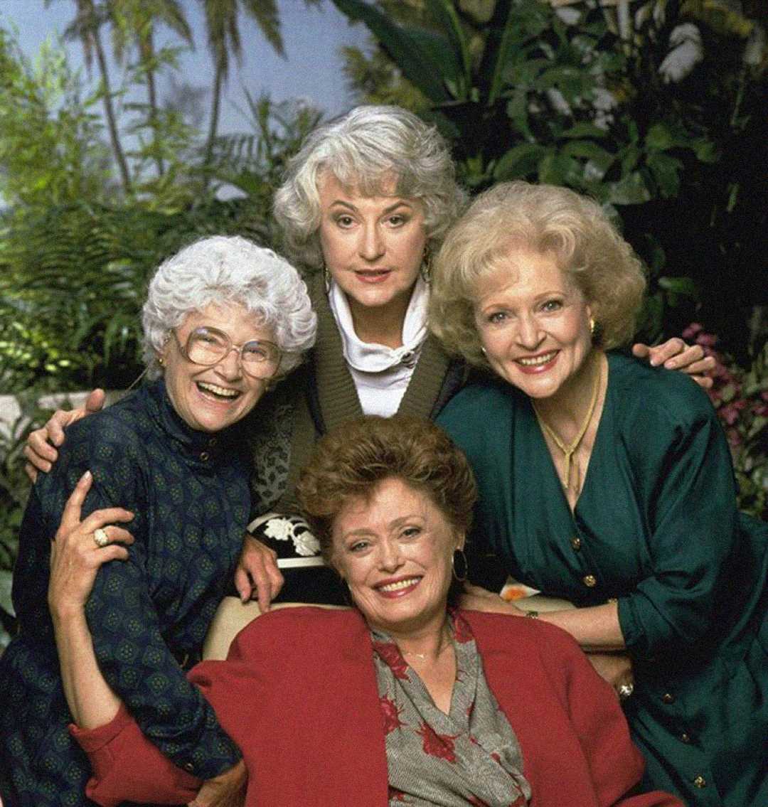 Four older women smile and pose together outdoors in front of lush greenery. Three women stand, while one sits in front. They wear colorful clothing and appear happy and friendly.