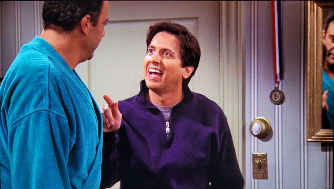 Two men stand facing each other indoors. The man on the left wears a teal robe, while the man on the right, in a dark purple sweatshirt, points and speaks animatedly. A medal hangs on the wall behind them.