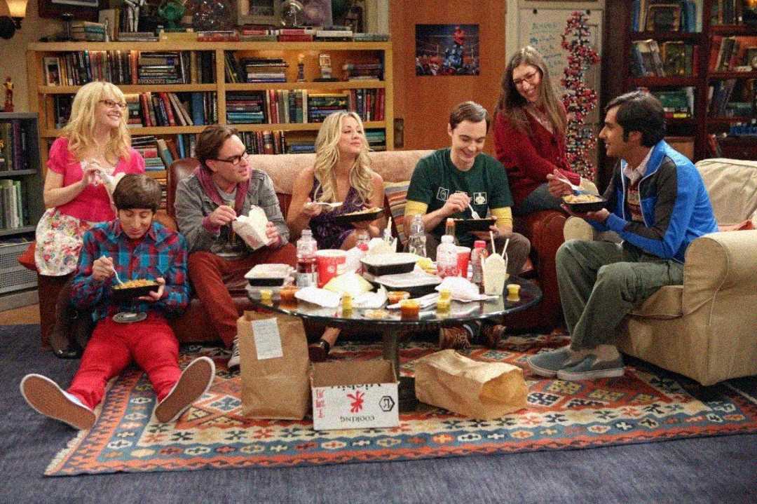 Seven people sit in a cozy living room, eating takeout food together around a coffee table covered with containers and drinks. They are smiling and engaged in conversation, with bookshelves and decorations in the background.