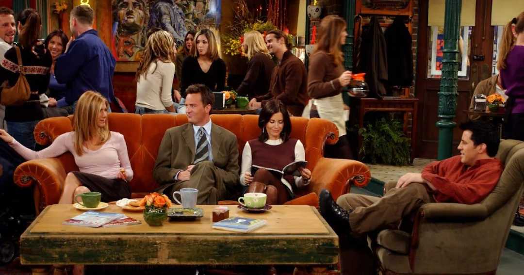 Four friends sit on a coffee shop couch and chair, chatting and relaxing with drinks. Other people mingle in the background, and the cozy shop is warmly decorated with plants and art.
