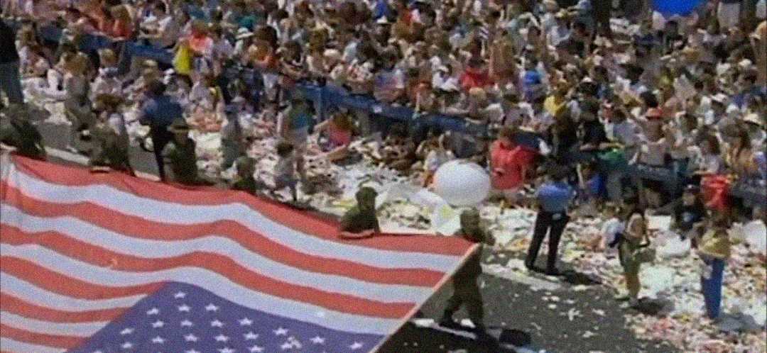 A large crowd watches a parade with people holding a giant American flag. Confetti and debris cover the street, and the audience sits behind a blue barrier.