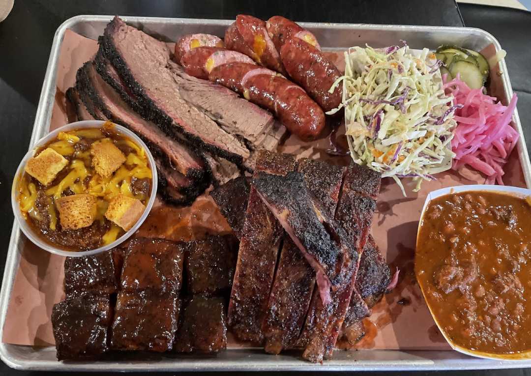 A tray with sliced brisket, sausage links, ribs, coleslaw, pickles, pickled onions, baked beans, mac and cheese with croutons, and glazed barbecue burnt ends.