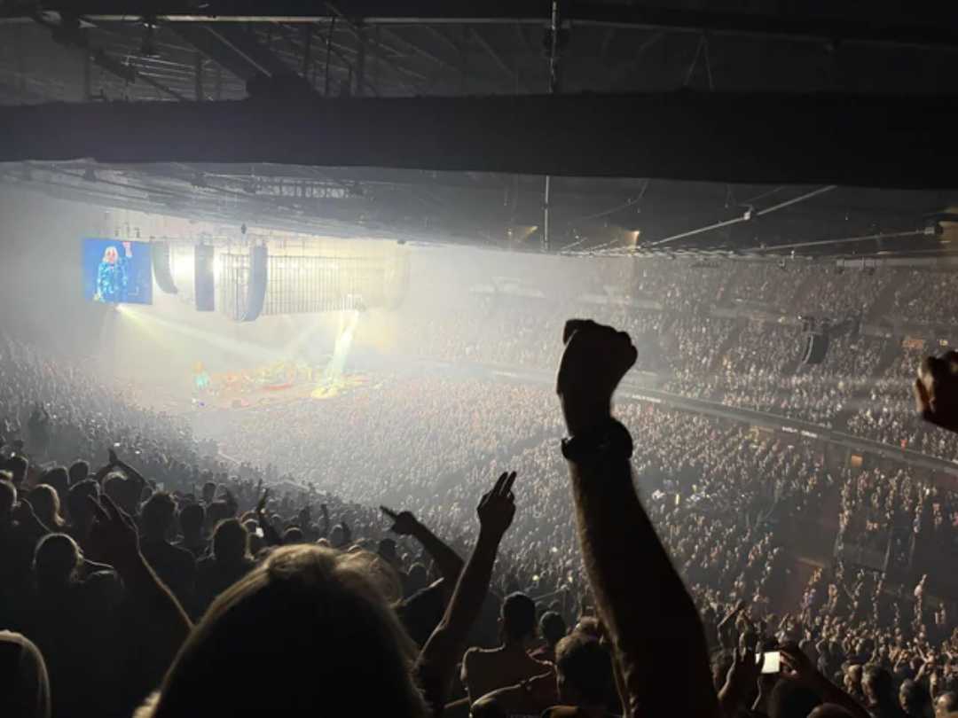 A large crowd fills an indoor arena, cheering with raised hands during a concert. Bright stage lights illuminate the performers, and screens display the show for the audience.