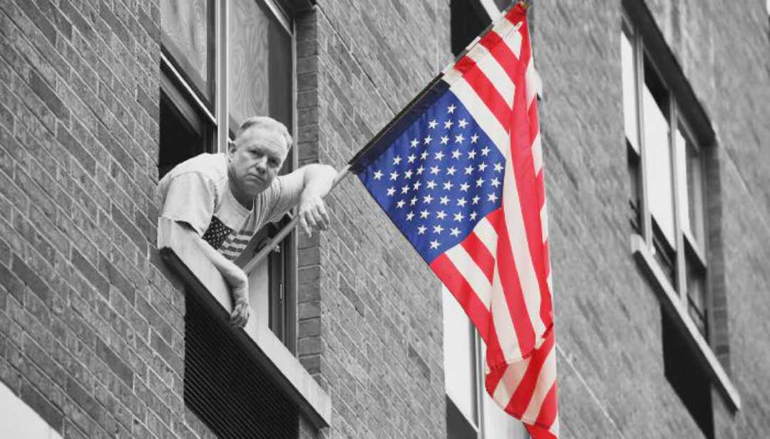 A man leans out of a window of a brick building, looking down with a serious expression. An American flag hangs outside the window, its colors vivid against the otherwise muted tones of the image.