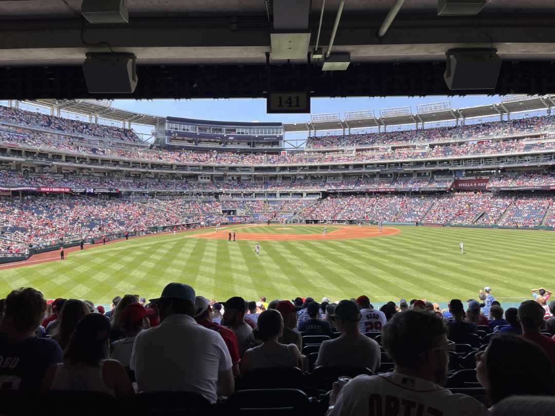 View from shaded stadium seats looking out onto a sunny baseball field during a game, with fans in the stands and players positioned on the field. The scoreboard and upper decks are visible in the background.