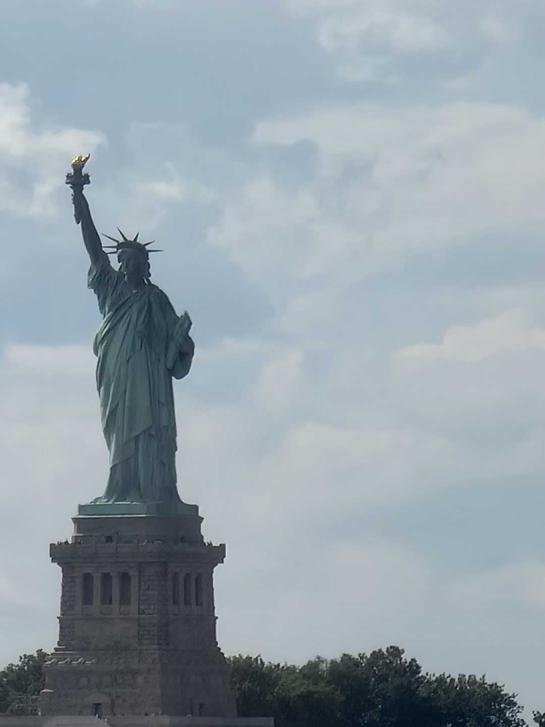 The Statue of Liberty stands tall on its pedestal against a partly cloudy sky, holding a torch in her right hand, with trees visible at the base of the structure.