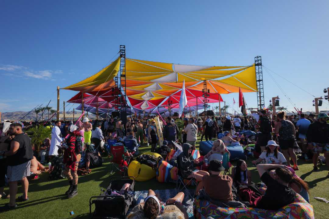 A large crowd gathers under a colorful, geometric canopy at an outdoor festival, with people sitting and standing on the grass, surrounded by bean bags and camping chairs on a sunny day.