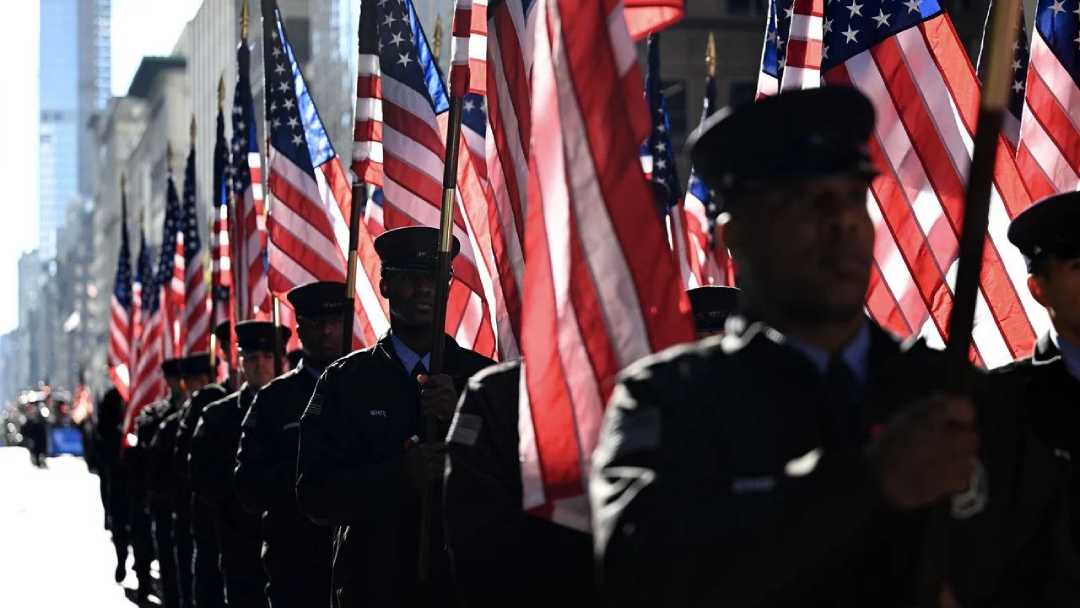 A group of uniformed individuals march in formation, each holding an American flag, with sunlight casting shadows across the street during a parade or ceremony.