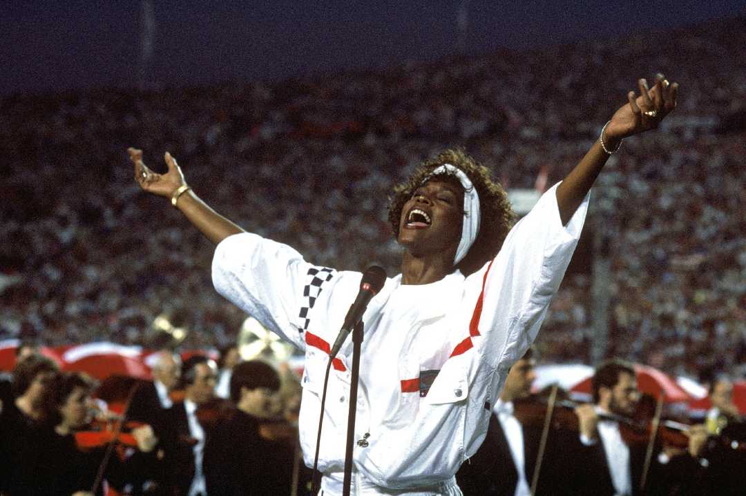A singer in a white outfit with arms outstretched performs passionately on a stadium field, with a microphone and orchestra behind them and a large crowd in the background.