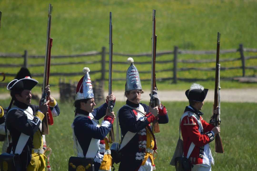 Four people in historical military uniforms stand outdoors, aiming muskets upward. They wear various hats and sashes, with a wooden fence and green field in the background. The scene appears to be a reenactment.