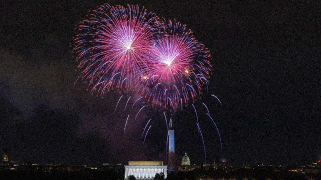 Bright fireworks burst in the night sky above the Washington Monument, with the Lincoln Memorial and the U.S. Capitol building visible below, illuminating the scene in vibrant colors.