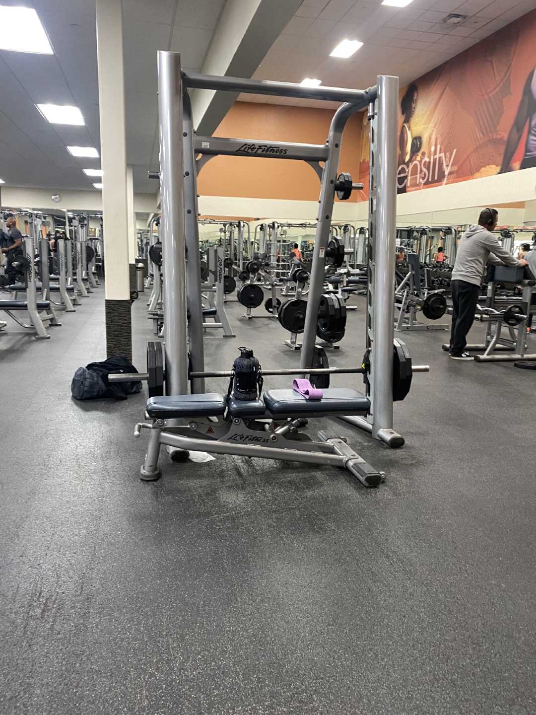 A gym with various exercise equipment, focusing on a Smith machine with weights, a water bottle, and a towel on it. People are working out in the background, and mirrors line the back wall.