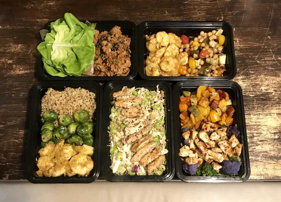 Six black meal prep containers on a wooden surface, each filled with portions of protein, vegetables, and grains, including grilled chicken, brown rice, brussels sprouts, lettuce, cauliflower, and mixed roasted vegetables.