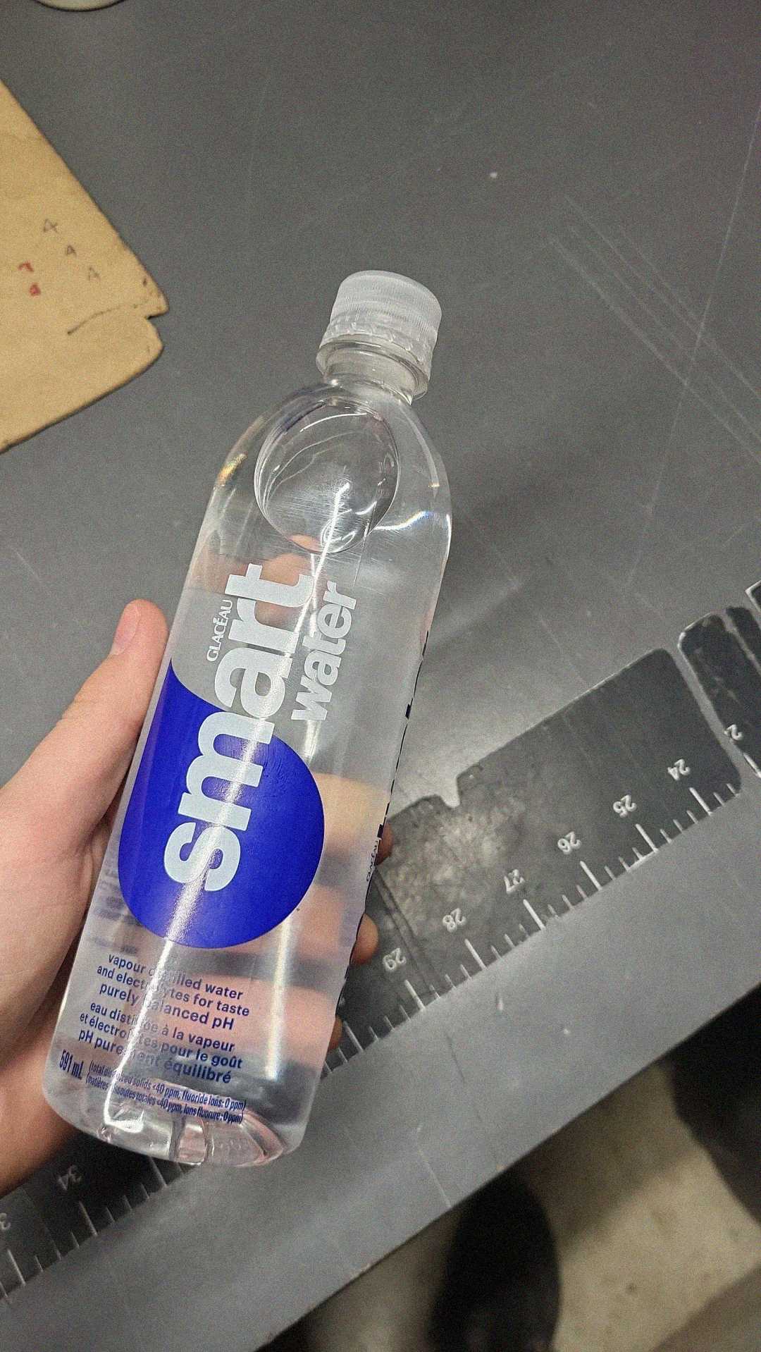 A hand holds a clear plastic bottle of Smartwater with a blue label over a gray table, next to a black ruler featuring white measurement markings.