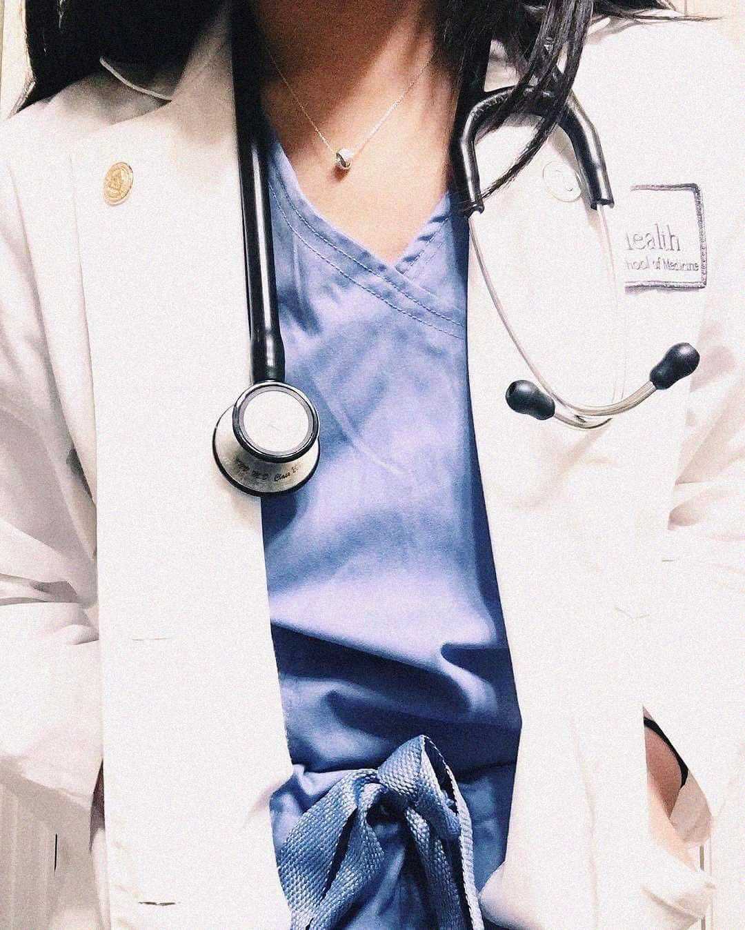 A person wearing a white lab coat and blue scrubs with a stethoscope draped around their neck, suggesting they are a medical professional.
